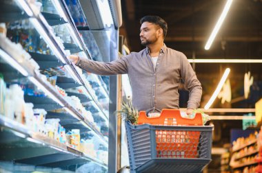 Parlak, modern bir markette soğutuculu raftan yiyecek seçen genç bir adam, el arabasını itiyor.