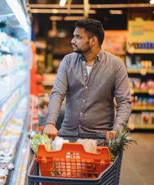 Markette alışveriş yapan, marul ve ananas gibi taze ürünlerle dolu bir arabayı iten, raflara bakan genç bir adam.