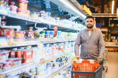 Süpermarkette alışveriş yapan bir adam, yiyecek dolu bir arabayı mandıra ürünlerinin yanından geçirir.