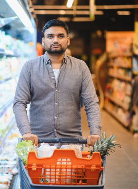 Parlak bir markette ananas ve marul gibi taze ürünlerle dolu bir alışveriş arabasını iten adam.