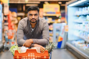 Süpermarkette alışveriş yapan genç bir adam. Taze meyve ve sebzelerle dolu bir alışveriş arabasına yaslanıyor.