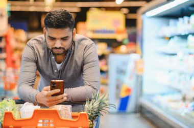 Elinde cep telefonu olan bir adam markette alışveriş arabasına yaslanmış dijital listesini kontrol ediyor.