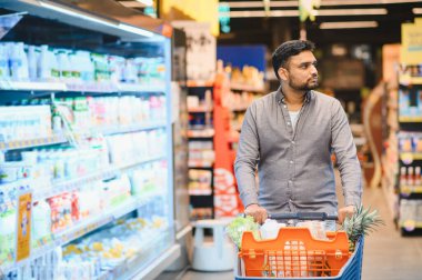 Genç adam market alışverişi yapıyor, modern bir süpermarketin mandıra ve içecek reyonunda dolu bir arabayı itiyor.