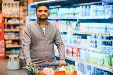 Genç bir adam, modern bir perakende süpermarkette yiyecek alırken taze ürünlerle market arabasını itiyor.