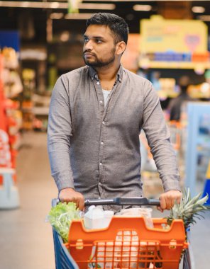Genç bir adam marketin koridorunda taze ürünlerle dolu bir alışveriş arabasını itiyor, raflarda satın alınacak eşyalar arıyor.