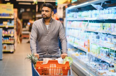 Genç bir adam, modern bir süpermarketin koridorunda içi yiyecek dolu bir alışveriş arabasını itiyor ve ürünleri seçmeye odaklanıyor.