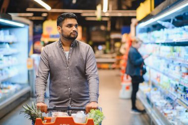 Adam soğutulmuş süpermarket koridorlarında kırmızı alışveriş arabalarını itiyor ürünleri tarıyor ve parlak modern mağazalardan taze sebzeler seçiyor.