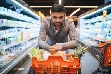 Adam makbuz ya da market listesini inceliyor. Parlak bir süpermarkette alışveriş arabasıyla geziyor. Taze yiyecek alıyor.