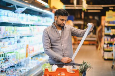 Adam elinde uzun bir market fişi tutuyor, markette alışveriş arabasını iterken eşyaları ve fiyatları dikkatle inceliyor.