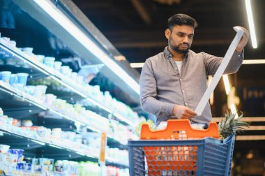Genç adam süpermarkette alışveriş yapıyor, alışveriş arabasındaki eşyalarla uzun bir market fişini kontrol ediyor. Enflasyon kavramı