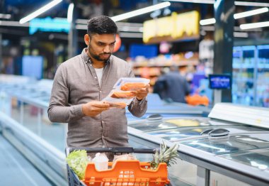 Yetişkin bir adam market alışverişi yapıyor, modern bir süpermarkette paketlenmiş balık tutuyor. Alışveriş arabası çeşitli taze ürünlerle dolu.