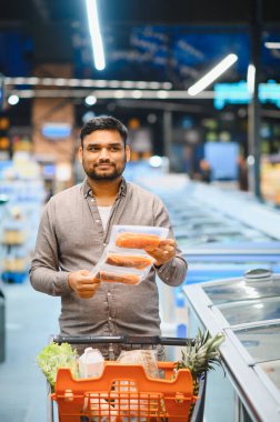 Market alışverişi yapan, buzdolabı koridorunun yanında vakumlu somon filetosu tutan sağlıklı yemek seçimleri yapan bir adam.