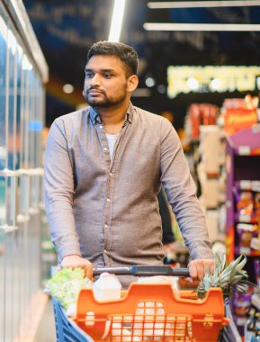 Taze ürünler ve temel ihtiyaçlarla dolu bir alışveriş arabasını iten adam, modern bir market koridorunda geziniyor.