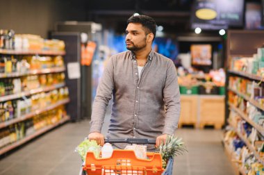 Market alışverişi yapan genç bir adam, taze ürünlerle dolu bir arabayı itmek ve parlak ışıklı bir süpermarket koridoru boyunca gerekli malzemeler.