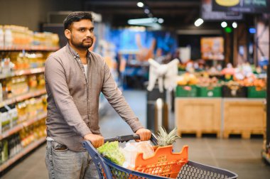 Taze ürünler ve paketlerle dolu bir alışveriş arabasını iten adam, parlak ışıklı bir market koridorunda yürüyor.