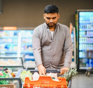 Perakende bakkal koridorunda içi taze meyve ve sebze dolu bir alışveriş arabasını iten genç adam.