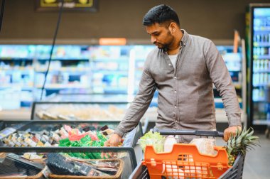 Genç bir adam alışveriş arabasını itiyor, modern bir süpermarkette dondurulmuş gıda vitrininden eşyalar seçiyor.