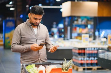Elinde akıllı telefon ve kağıt liste tutan bir adam perakende mağazasında market arabasını iterken eşyaları karşılaştırıyor.