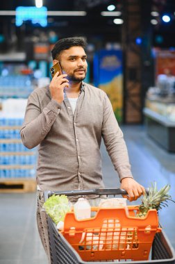 Alışveriş arabasını iten bir adam, market koridorunda bir telefon görüşmesi yapıyor.