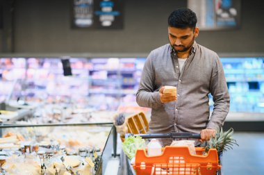 Elinde peynirle süpermarket koridorundaki ürünleri alışveriş arabasıyla karşılaştıran genç bir adam.