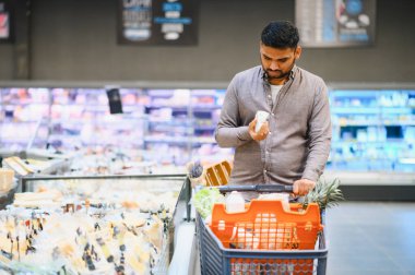 Hintli bir adam market alışverişi yapıyor, market reyonunda bir ürünü incelerken el arabasını itiyor.