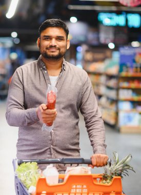 Genç adam markette alışveriş yapıyor, elinde paketlenmiş bir salam tutuyor ve taze ürünlerle dolu bir arabayı itiyor.