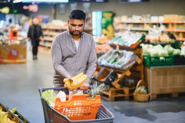 Modern bir süpermarkette alışveriş yaparken elinde muz tutan ve alışveriş arabasıyla alışveriş yapan genç bir adam.