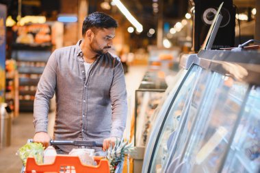 Alışveriş arabalı bir adam markete doğru gidiyor, taze ürünlere ve soğutulmuş bölümlere bakıyor.