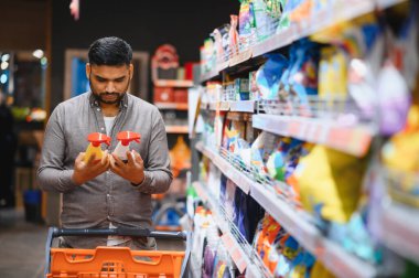Benzer iki sprey şişesi tutan bir adam markette ev eşyaları alırken ürünleri karşılaştırıyor.