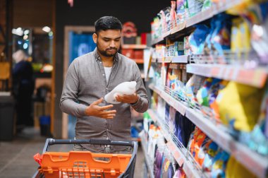Müşteri temel ihtiyaçlar için alışveriş yapıyor, bir ürün şişesini inceliyor ve raflarda bir el arabasını itiyor.