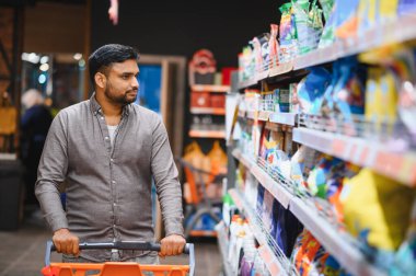 Adam boş bir alışveriş arabasını itiyor, market raflarındaki ürünlere bakıyor.