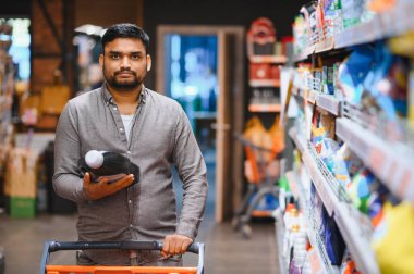Alışveriş arabalı bir adam markette elinde bir şişe sıvı deterjan tutarak ev için günlük ihtiyaçlarını karşılıyor.