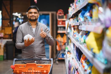 Mutlu adam temizlik malzemesi seçerken ve süpermarket koridorunda alışveriş arabası iterken, güvenle gülümserken başparmağını kaldırıyor.