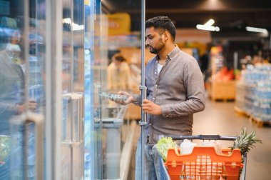 Modern bir süpermarket koridorunda alışveriş yaparken bir kutu yumurta seçmek için buzdolabının kapısını açan bir adam.