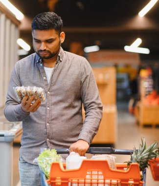 Elinde bıldırcın yumurtası olan bir adam markette içi yiyecek dolu bir alışveriş arabasını itiyor ve sağlıklı yiyecek seçimleri yapıyor.