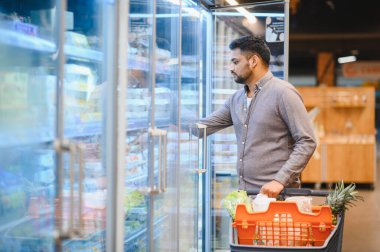 Dondurucu vitrinindeki ürünlere uzanan bir adam, içi yiyecek dolu bir alışveriş arabasını itiyor.