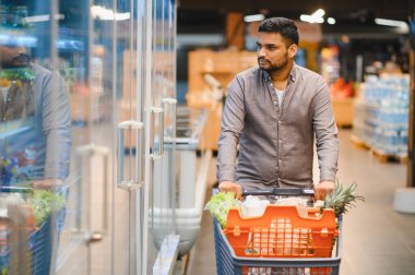 Erkek tüketici taze ürün ve bakkaliye alışverişi yapıyor, süpermarkette soğutulmuş bölümün önünden geçiyor.