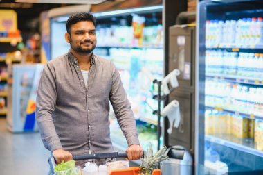 Genç bir adam, marketin canlı bir koridorunda içi yiyecek dolu bir alışveriş arabasını itip sağlıklı yiyecekleri seçiyor.