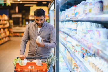 Alışveriş arabasını iten bir adam, bir süpermarketin mandıra koridorunda ürün etiketini dikkatle inceleyen, tüketici seçimi yapan bir adam.