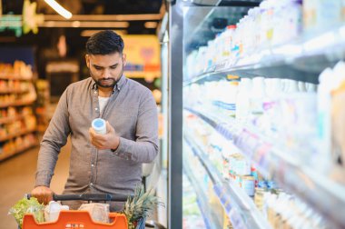 Bir ürünü elinde tutan, beslenme bilgilerini okuyan ve bir markette satın alma kararları veren bir adam.