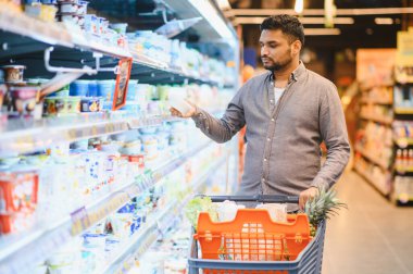 Genç adam bakkal alışverişi yapıyor. Parlak bir perakende mağazasındaki soğutuculu raftan yoğurt seçiyor.
