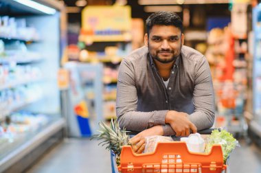 Genç Hintli bir adam süpermarket koridorunda içi taze ürünlerle dolu bir alışveriş arabasına yaslanmış kameraya gülümsüyor.