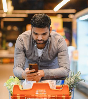 Modern bir süpermarkette yiyecek dolu bir market arabasına yaslanırken akıllı telefondan alışveriş listesini kontrol eden adam.