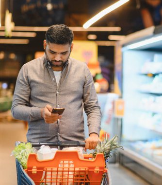 Market koridorunda gezinen genç bir adam alışveriş listesi için akıllı telefon ekranına odaklanırken alışveriş arabasını itiyor.