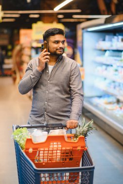 Alışveriş arabasını iten, market alışverişi yapan, telefon eden, bakkal koridorunda gezinen bir adam.