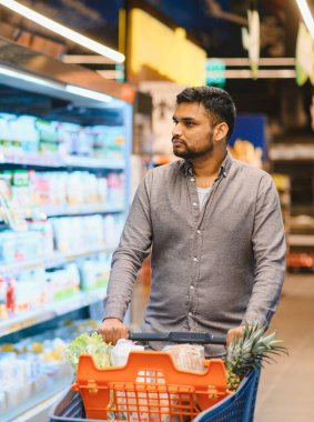Süpermarket koridorunda içi taze ürünlerle dolu bir alışveriş arabasını iten adam evi için eşyalar seçiyor.