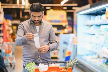Bir süpermarkette uzun bir market fişine bakan adam, yükselen fiyatlar ve artan yaşam maliyeti hakkında endişeli.