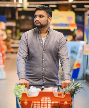 Genç adam taze ürünler ve gerekli malzemeler için alışveriş yapıyor, market koridorunda bir el arabasını itiyor.