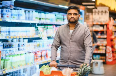 Genç, yetişkin bir adam markette içi yiyecek dolu bir alışveriş arabasını itiyor. Parlak bir süpermarket koridorunda ürünleri tarıyor.