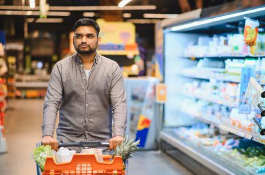 Genç adam modern süpermarket koridorlarında turuncu alışveriş arabalarını itiyor, taze ürünleri ve bakkaliyeleri seçiyor.